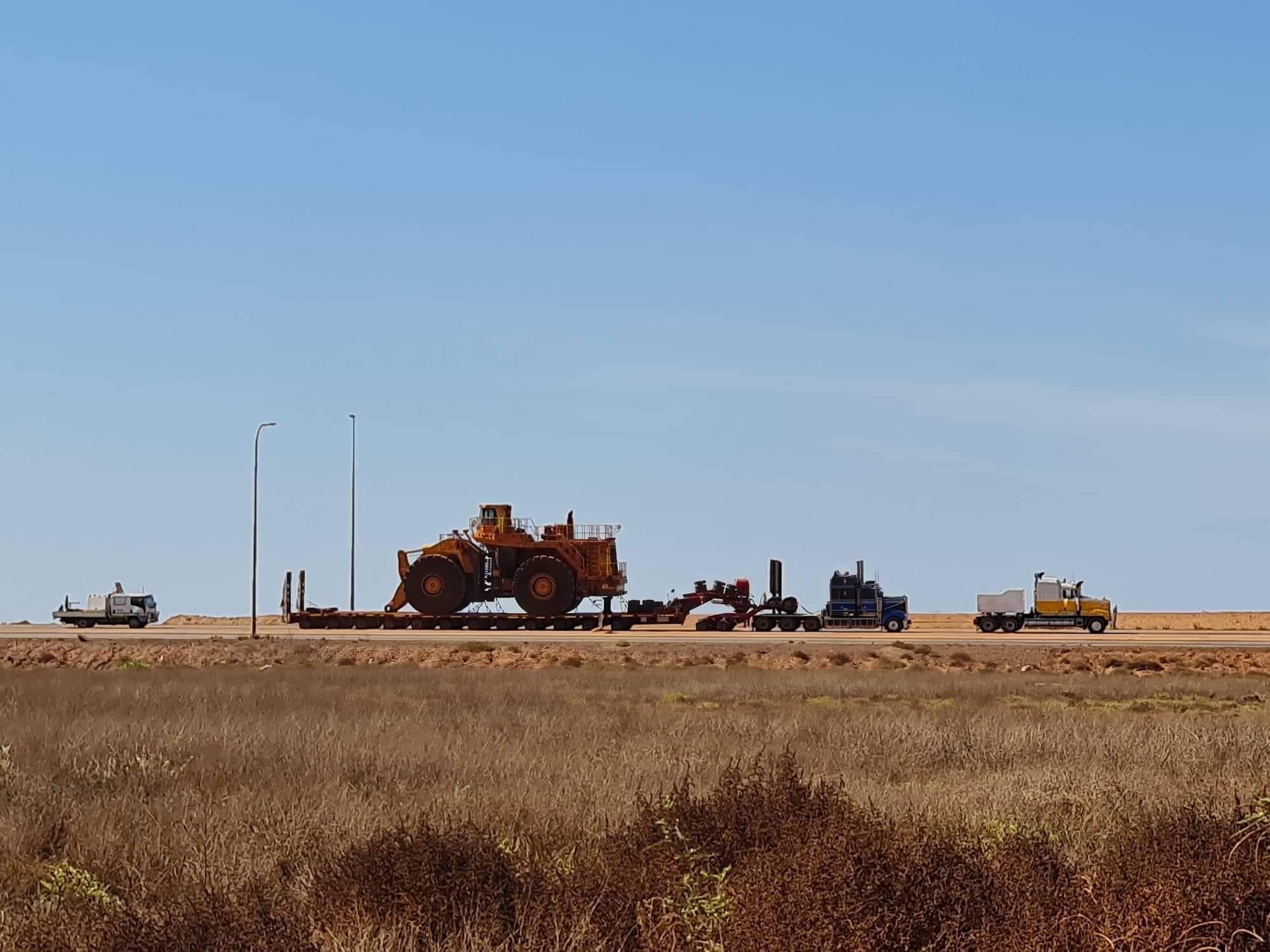Side view of a heavy vehicle convoy featuring three trucks, one with a long trailer towing a earthmoving machine, moving along a road